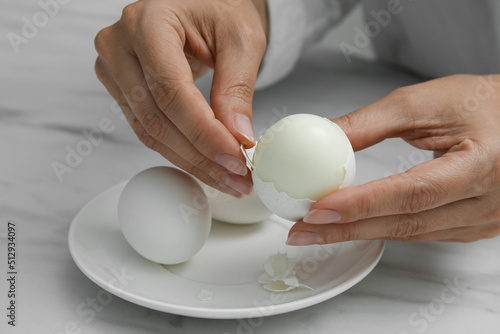 Woman peeling boiled egg at white marble table, closeup photo