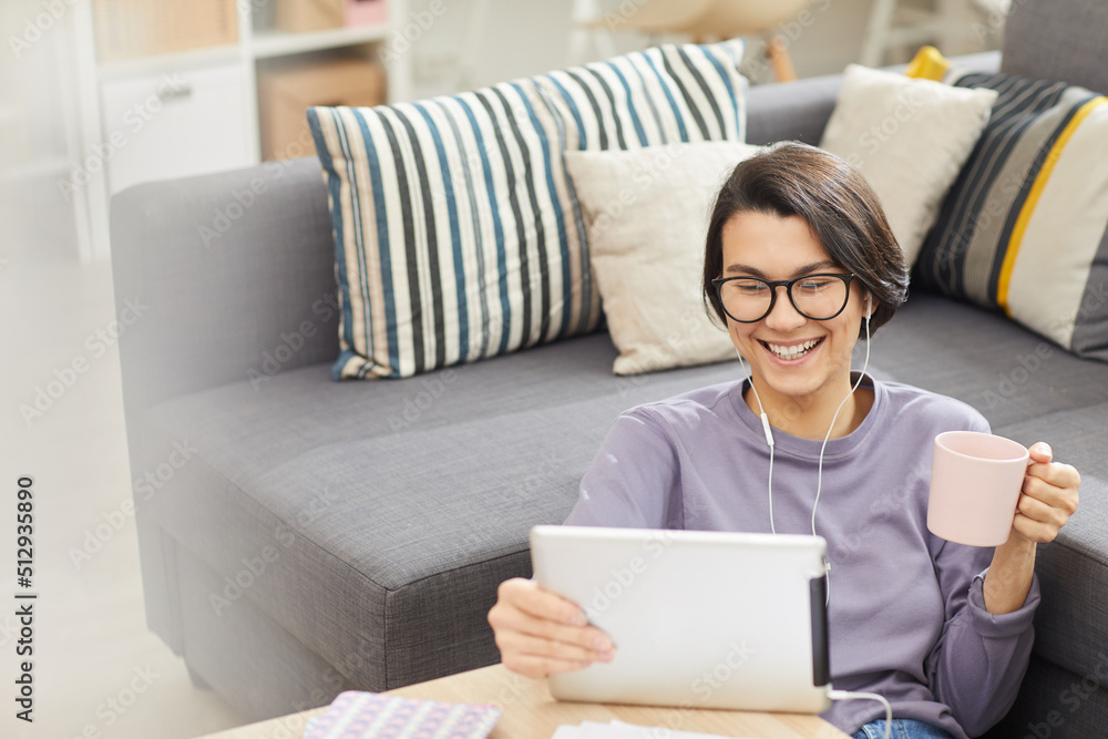 Excited young brunette woman in glasses leaning on sofa and drinking coffee while making video call via tablet app Excited young brunette woman in glasses leaning on sofa and drinking coffee while making video call via tablet app