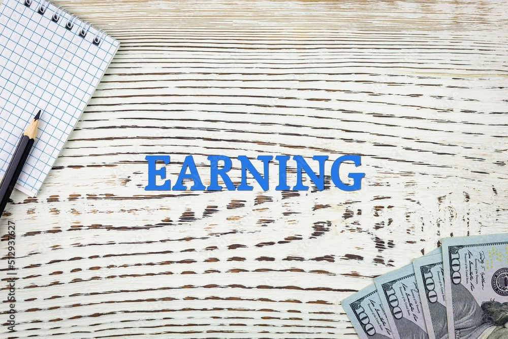 Naklejka premium EARNING - word (text) and dollars on a white wooden table, notebook, notepad. Business concept: buying, selling, commerce (copy space).