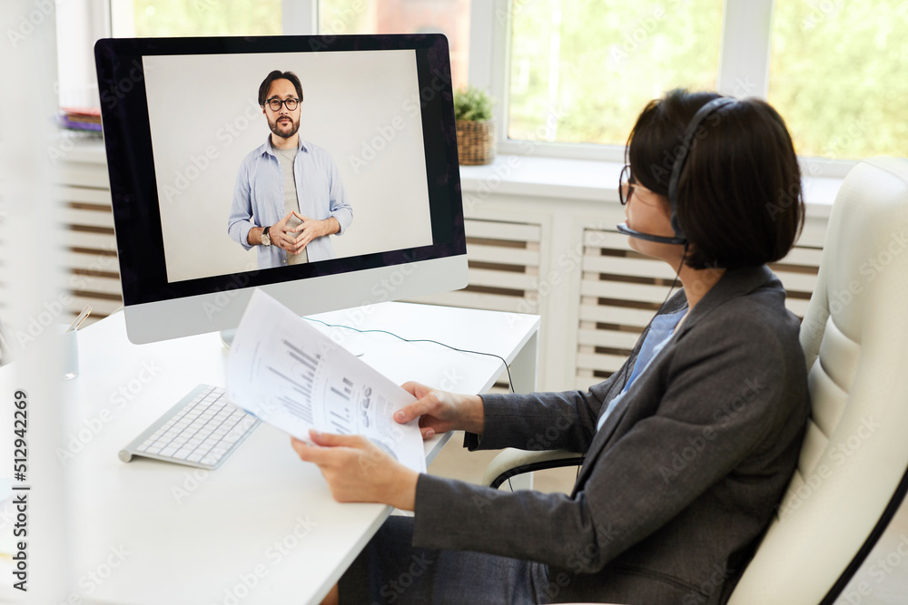 Serious young business analyst sitting at desk in office and analyzing statistics while interacting with Asian colleague via web conferencing provider Serious young business analyst sitting at desk in office and analyzing statistics while interacting with Asian colleague via web conferencing provider