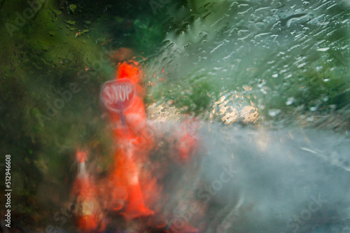 Worker wearing high visibility orange clothing with Stop sign in the heavy rain. Photo taken using intentional camera movement behind rain spattered car window.