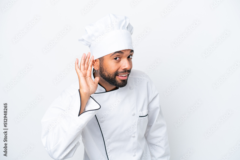 Young Brazilian chef man isolated on white background listening to something by putting hand on the ear Young Brazilian chef man isolated on white background listening to something by putting hand on the ear