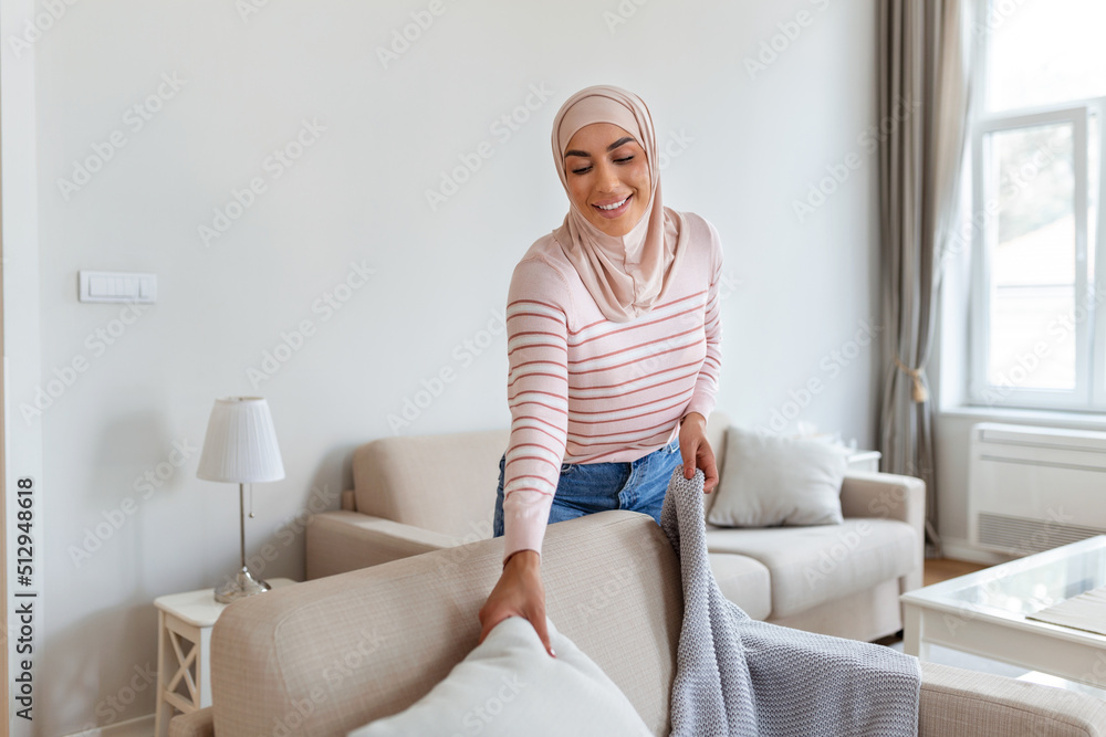 Lovely young Arabic Muslim woman putting soft pillows and plaid on comfy sofa, making her home cozy and warm Lovely young Arabic Muslim woman putting soft pillows and plaid on comfy sofa, making her home cozy and warm