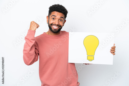 Young Brazilian man isolated on white background holding a placard with bulb icon and celebrating a victory