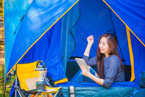 Beautiful lady wearing a blue shiry and jean sitting inside a tent, she holding her notepad and the other hand holding a pencil at the camping area. yellow cahir with lantern. happy face, smiling face photo