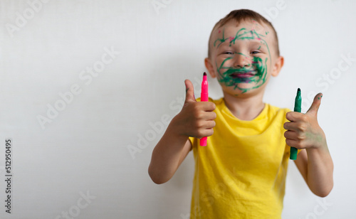 A 4-year-old boy painted his face with a marker. The boy stands against a white wall, holding markers in his hands, smiling and showing the class. Banner with space for text