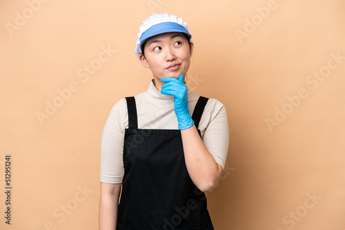 Young Chinese Fishmonger woman wearing an apron and holding a raw fish isolated on pink background and looking up