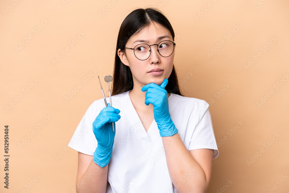 Dentist Chinese woman holding tools isolated on beige background having doubts and thinking Dentist Chinese woman holding tools isolated on beige background having doubts and thinking