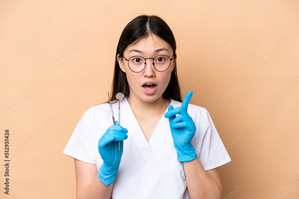 Dentist Chinese woman holding tools isolated on beige background intending to realizes the solution while lifting a finger up Dentist Chinese woman holding tools isolated on beige background intending to realizes the solution while lifting a finger up