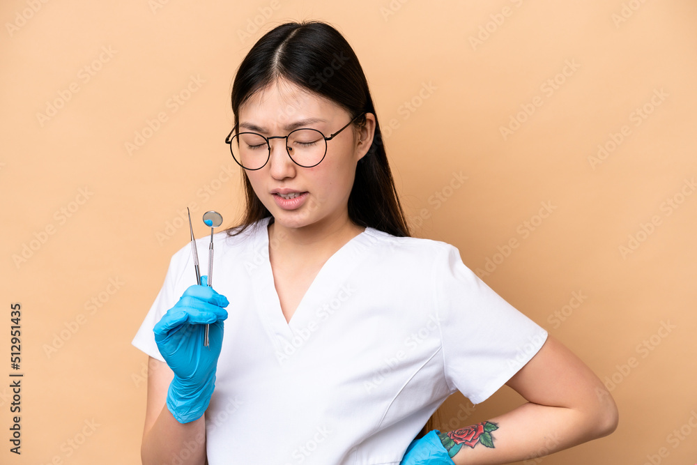 Dentist Chinese woman holding tools isolated on beige background suffering from backache for having made an effort Dentist Chinese woman holding tools isolated on beige background suffering from backache for having made an effort