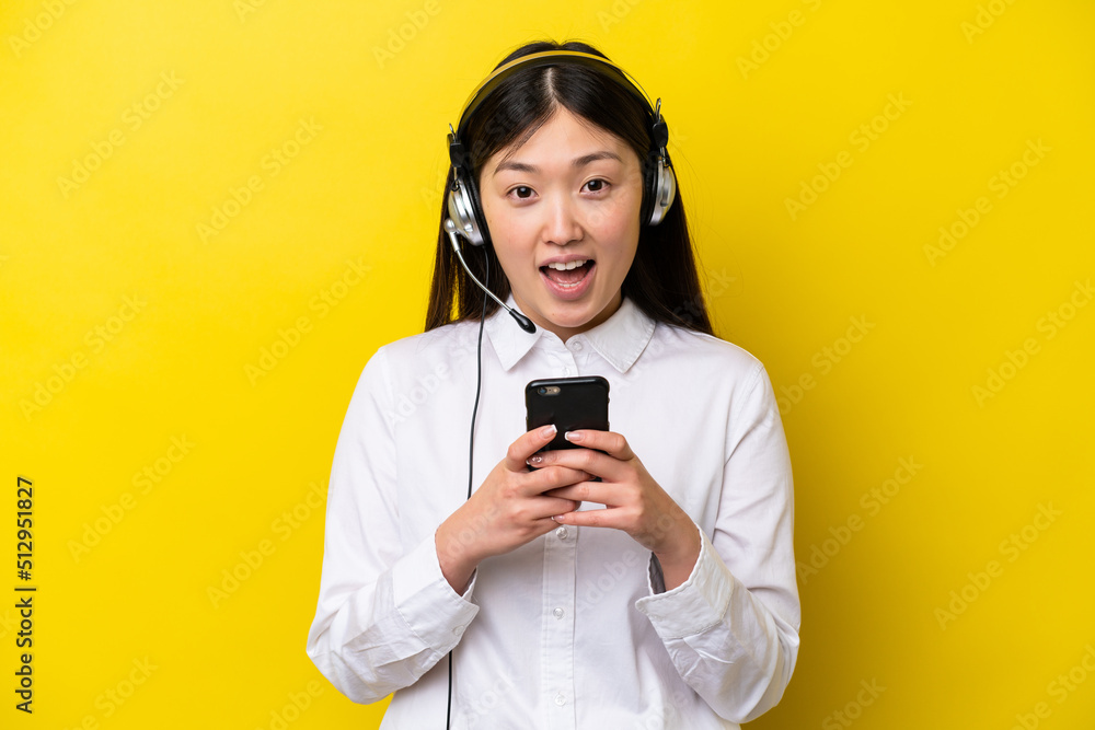Telemarketer Chinese woman working with a headset isolated on yellow background surprised and sending a message Telemarketer Chinese woman working with a headset isolated on yellow background surprised and sending a message
