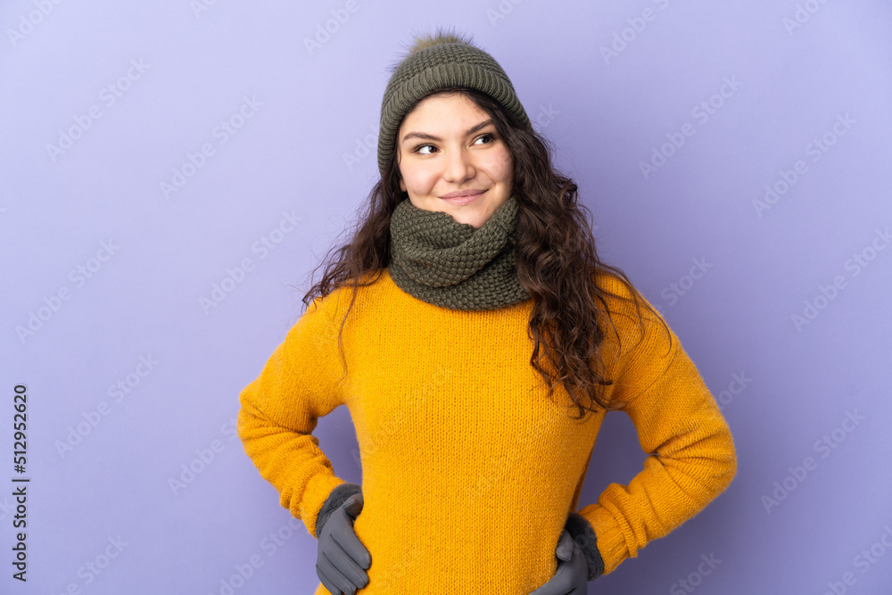 Teenager Russian girl with winter hat isolated on purple background posing with arms at hip and smiling Teenager Russian girl with winter hat isolated on purple background posing with arms at hip and smiling