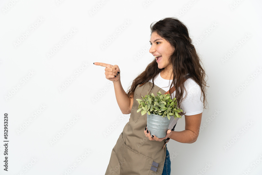 Young Russian gardener girl holding a plant isolated pointing finger to the side and presenting a product Young Russian gardener girl holding a plant isolated pointing finger to the side and presenting a product
