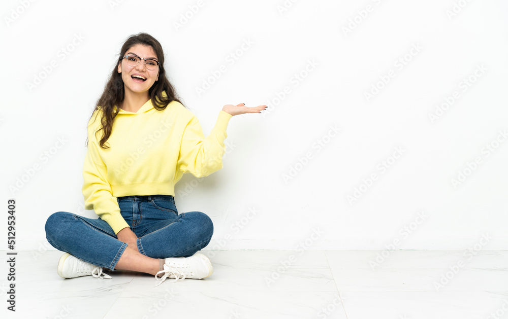 Teenager Russian girl sitting on the floor holding copyspace imaginary on the palm to insert an ad Teenager Russian girl sitting on the floor holding copyspace imaginary on the palm to insert an ad