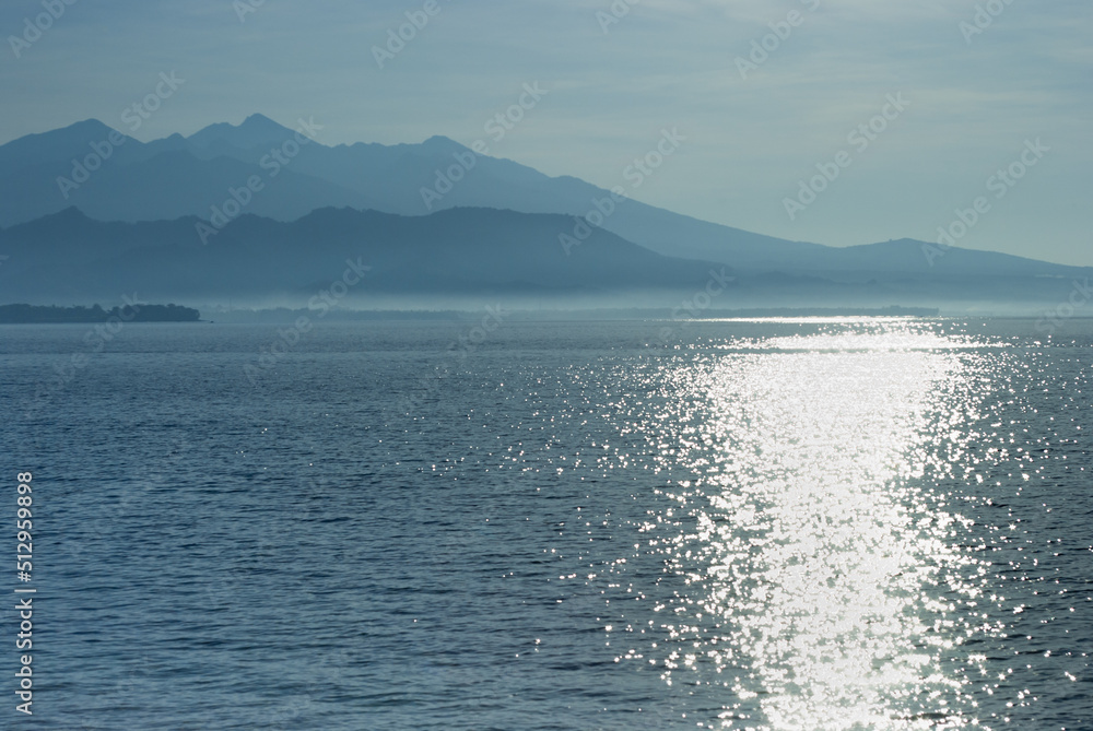 Bright shiny sunrise on sea with blue mountains in haze on horizon, sunny calm sea and sun glares on water. Fresh sunny morning on indonesian island, summer travel on asia.