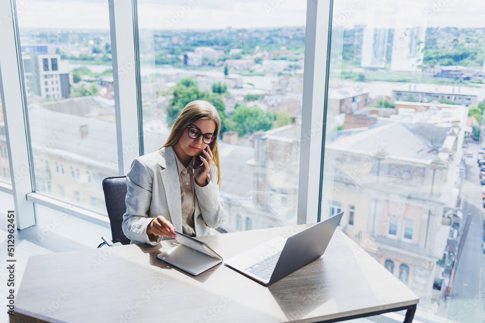 A fair-haired cheerful European woman in glasses in stylish casual clothes is sitting at a table with a laptop, doing paperwork and talking on the phone. Business lady at the workplace in the office