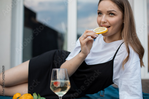 Happy young woman lying on blue plaid on terrace with glass of white wine and eating orange closeup. Drinking alcohol drink and enjoying outdoor recreation. Home party, vacation and lounge