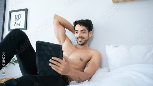 Fotografi Muscular Hispanic man lying in bed under a black blanket with smartphone in hands