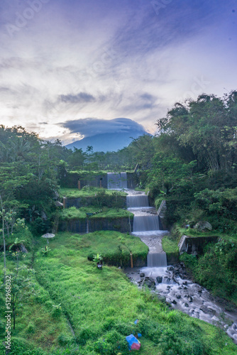 Merapi volcano with lenticular cloud on the peak of mountain with terraced waterfall on the foreground and sunrise sky in slightly foggy weather. View from Mangunsuko or jokowi bridge, Indonesia photo