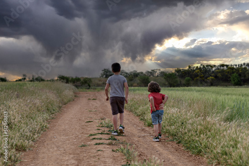 Two Caucasian boys walk through the field looking at a storm in the sky.