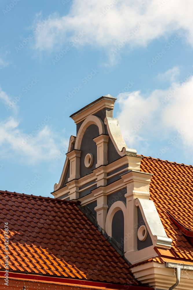 Vertical photo of red metal roofs and grey white baroque roof decoration at blue sky background. A shadow fell to roof from decoration in Kaunas, Lithuania, Europe