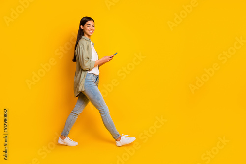 Full length profile photo of pretty positive girl hold telephone walk empty space isolated on yellow color background