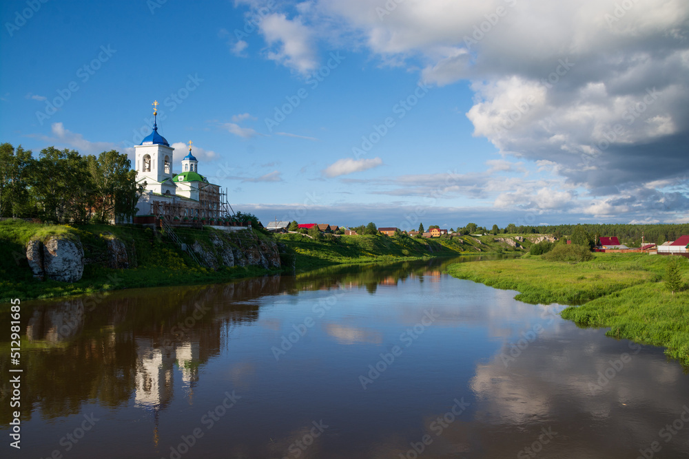 an old Orthodox church and a village on the river bank.sun , blue sky and black clouds.