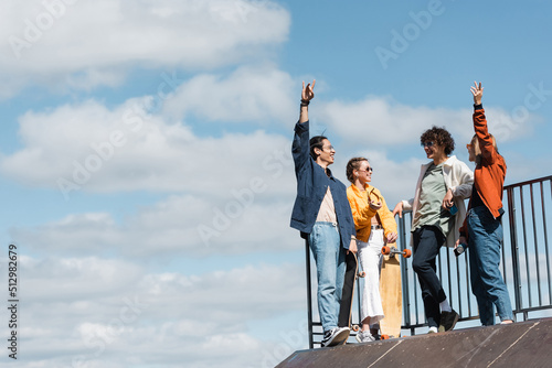 cheerful interracial friends gesturing on skate ramp under blue and cloudy sky.