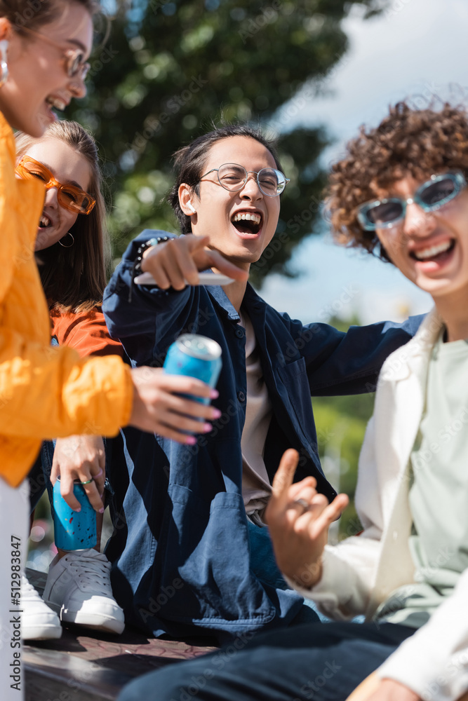 excited asian man in eyeglasses pointing with finger near blurred friends outdoors. excited asian man in eyeglasses pointing with finger near blurred friends outdoors.