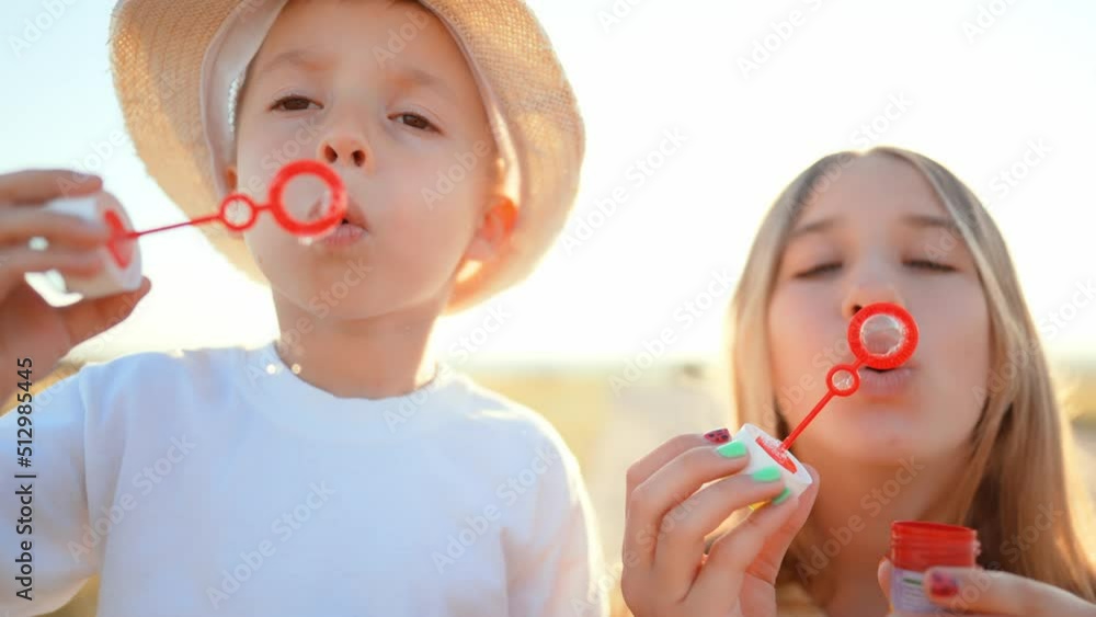 Teenage sister stands and smiles next to little brother who blows and gets soap bubbles in outdoor in field. Family walk on weekend out of town in countryside. Leisure activity. Healthy lifestyle.