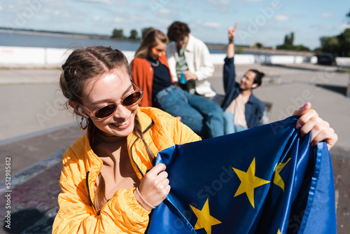 Фотография happy woman in earphone and sunglasses holding eu flag near blurred friends