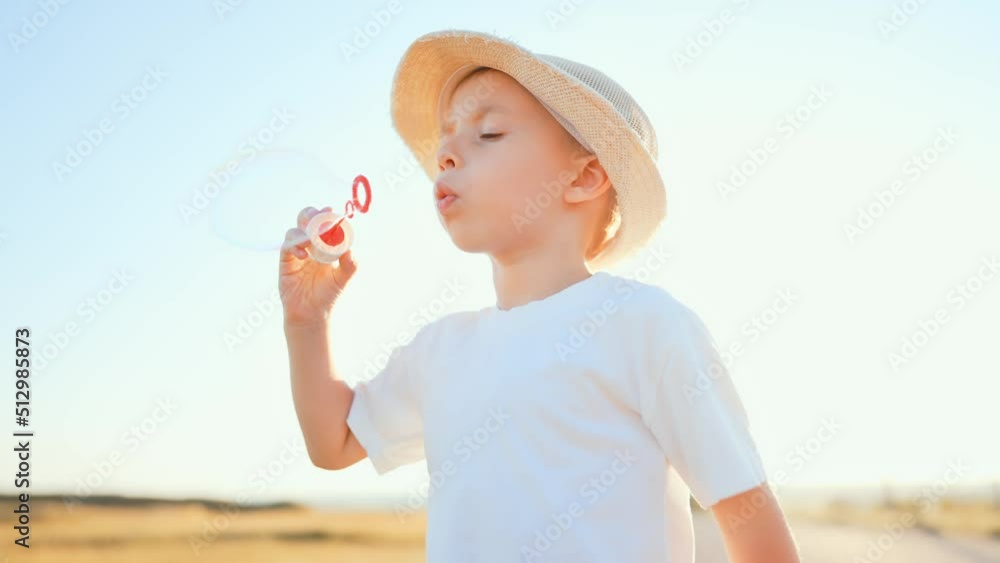 Close up portrait baby boy stands in straw hat on country road in field in summer under the sun rays and blows soap bubbles with rainbow reflection. Games in childhood outdoor on leisure activity. Close up portrait baby boy stands in straw hat on country road in field in summer under the sun rays and blows soap bubbles with rainbow reflection. Games in childhood outdoor on leisure activity.