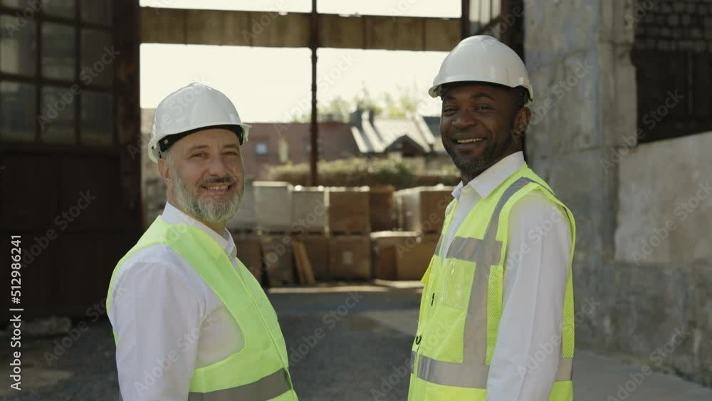 Group of two civil engineers smiling and looking at camera while standing opposite to each other. Multicultural coworkers cooperating together for realization industry project at construction site. Group of two civil engineers smiling and looking at camera while standing opposite to each other. Multicultural coworkers cooperating together for realization industry project at construction site.