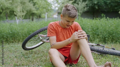 The young handsome boy is crying and upset that he fell off the bike and got painful leg injury. The child is holding the knee because the wound on the skin hurts. Accident with bicycle in green park