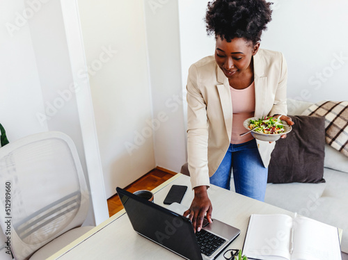Portrait of a young African American businesswoman eating lunch in an office. Mid adult African business woman eating salad for lunch while working on her laptop in her home office.