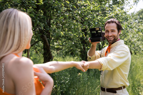 Fashionable man holding retro camera and holding hand of blurred girlfriend in park.
