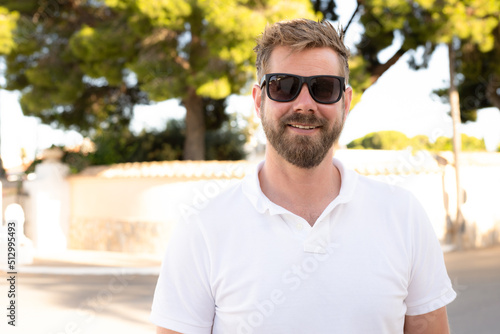 Outdoor shot of beautiful bearded young man in sunglasses posing over city garden on warm sunny day, wearing white polo shirt