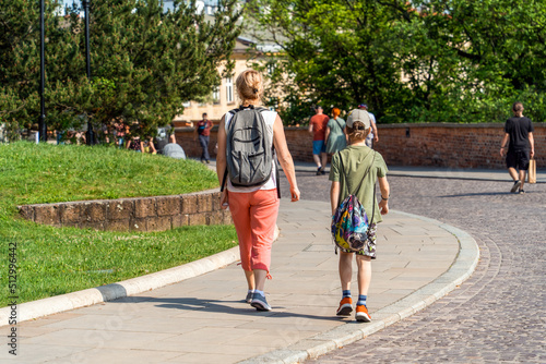 Mom and teenage son with backpacks behind their backs in summer clothes on a sunny day walk along the sidewalk next to the green lawn in the old town among passers-by, family travel © Oleksandr Bochkala