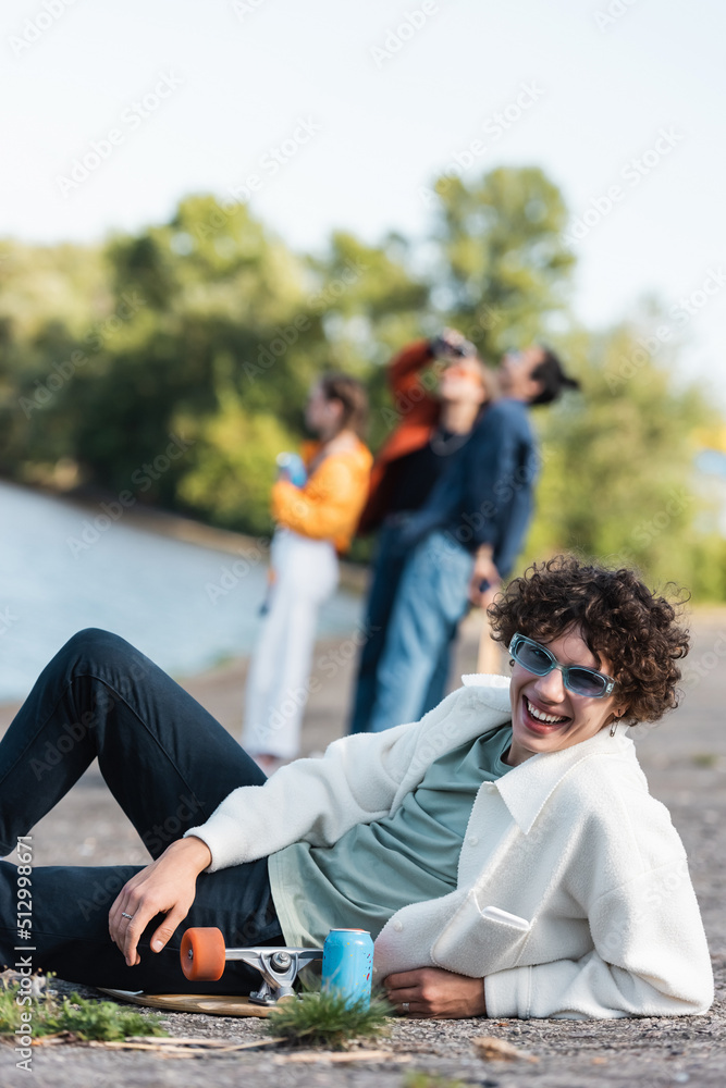 curly man in trendy sunglasses smiling at camera near skateboard and friends on blurred background. curly man in trendy sunglasses smiling at camera near skateboard and friends on blurred background.
