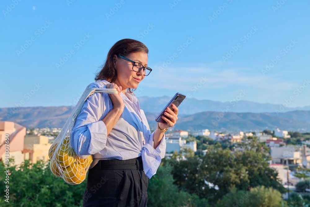 Outdoor mature woman with smartphone bag of oranges in mountain countryside