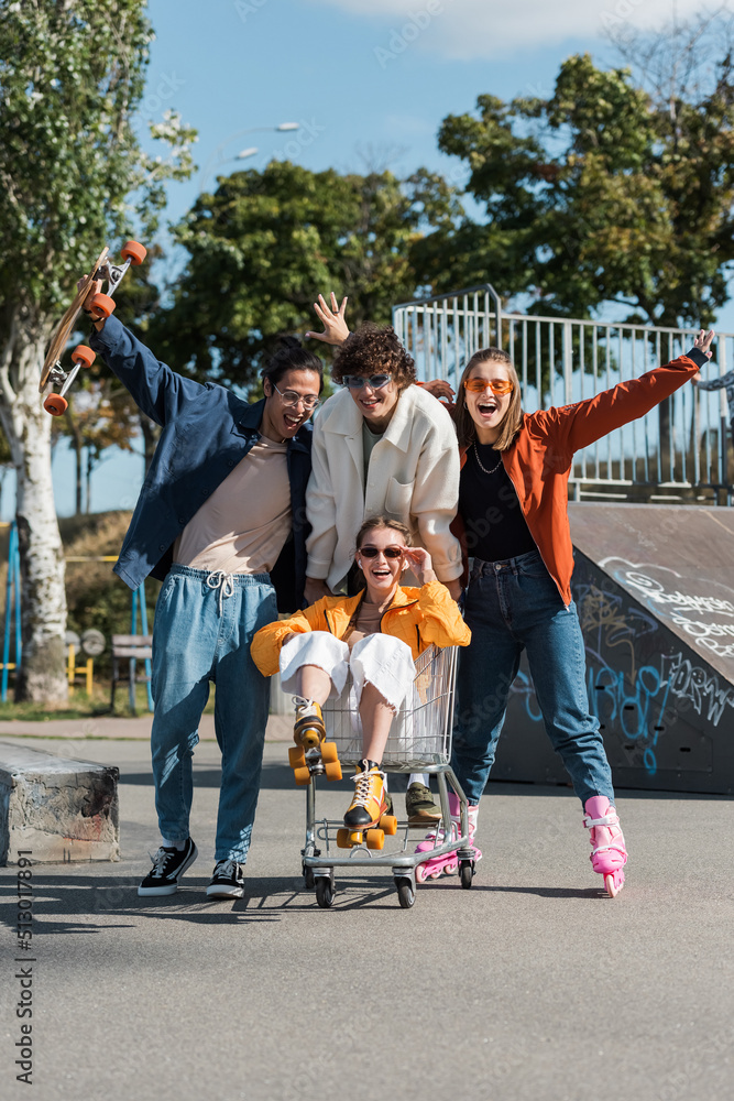 Obraz premium excited multiethnic skaters moving shopping cart with happy woman in skate park.