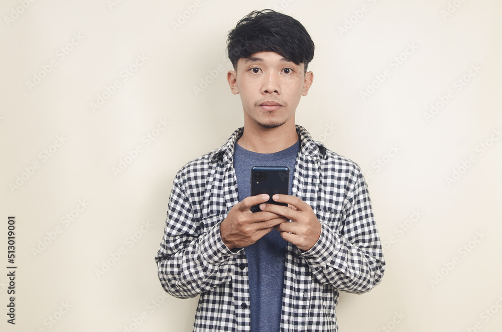 Close-up portrait of young Asian male model wearing plaid shirt while holding phone on isolated background Close-up portrait of young Asian male model wearing plaid shirt while holding phone on isolated background