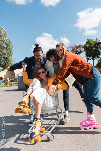 woman in rollers skates waving hand in shopping cart near cheerful multiethnic friends.
