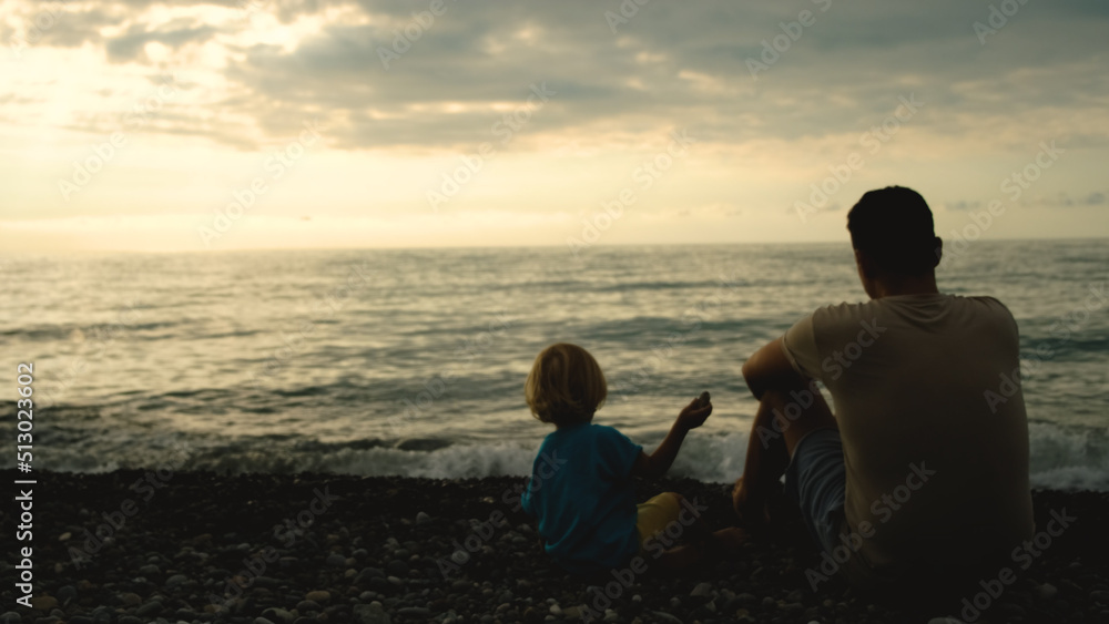 Father and son sit back on the beach and throw stones into the sea at the sunset time. Concept of friendly family.
