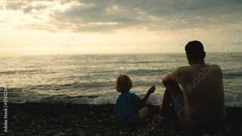 Father and son sit back on the beach and throw stones into the sea at the sunset time. Concept of friendly family. © Voyagery