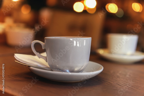 White tea cup standing on saucer in soft focus on naturally blurred background with bokeh lights. Coffee cup on rustic wooden table. Coffee, tea house. The concept of a cozy pastime, tea ceremony