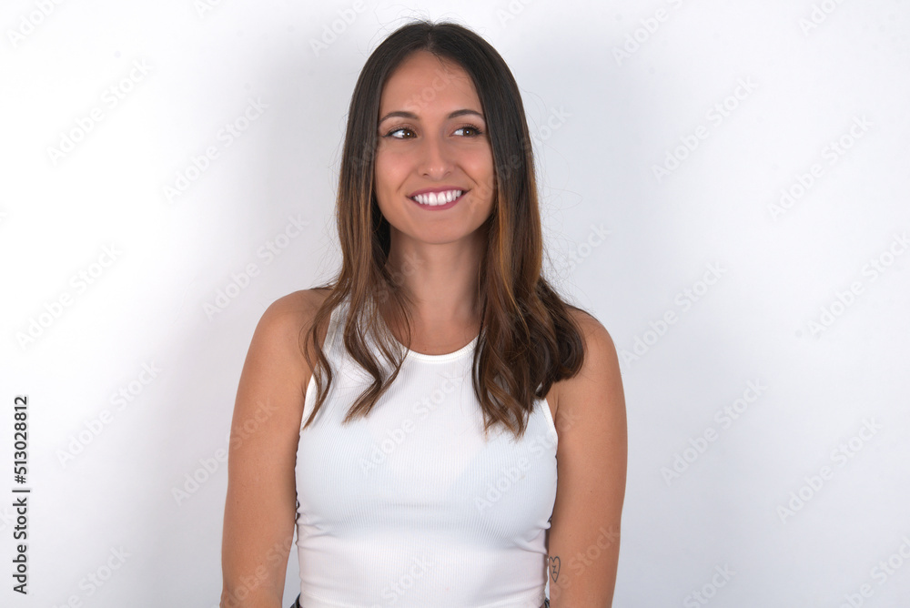 young beautiful caucasian woman wearing white top over white background with thoughtful expression, looks away keeps hands down bitting his lip thinks about something pleasant. young beautiful caucasian woman wearing white top over white background with thoughtful expression, looks away keeps hands down bitting his lip thinks about something pleasant.