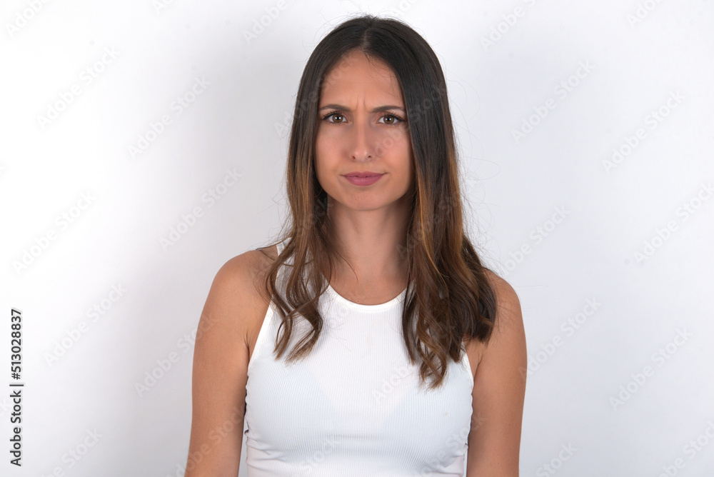 young beautiful caucasian woman wearing white top over white background frowning his eyebrows being displeased with something. young beautiful caucasian woman wearing white top over white background frowning his eyebrows being displeased with something.