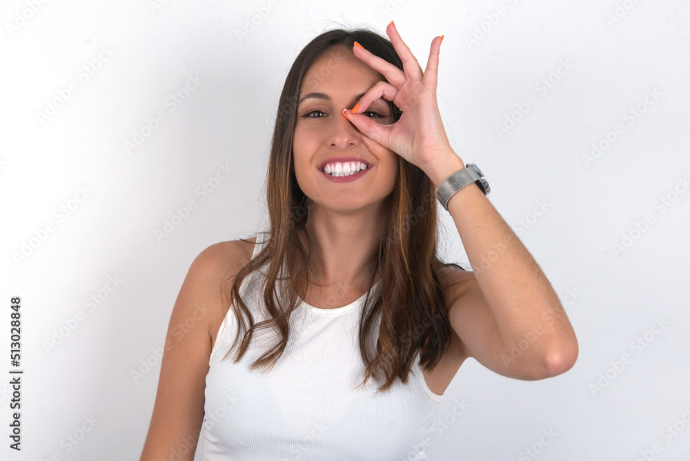 young beautiful caucasian woman wearing white top over white background with happy face smiling doing ok sign with hand on eye looking through finger. young beautiful caucasian woman wearing white top over white background with happy face smiling doing ok sign with hand on eye looking through finger.