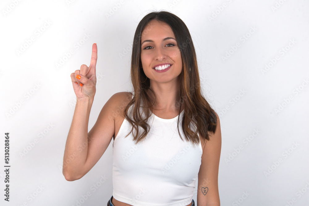 young beautiful caucasian woman wearing white top over white background showing and pointing up with fingers number one while smiling confident and happy. young beautiful caucasian woman wearing white top over white background showing and pointing up with fingers number one while smiling confident and happy.